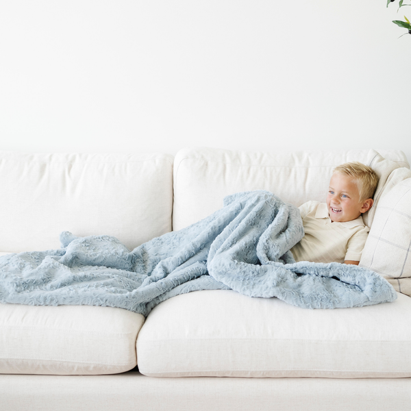 Boy lays on couch snuggled in a blue cozy faux fur toddler blanket. 