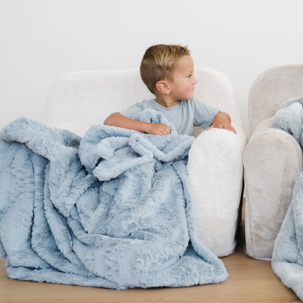 Little boy sits in a cozy chair with a blue faux fur soft toddler blanket. 
