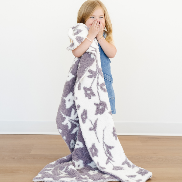 Little girl stands snuggled with a purple and cream floral double layer bamboni toddler blanket. 