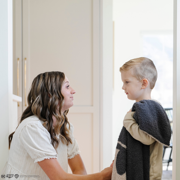 A little boy stands by his mom while holding a black and light gray double-layer Bamboni® blanket with a Batman™ icon on it. The soft blanket is a luxury blanket and a kids blanket as well as a Saranoni blanket. This kids blanket is perfect for littles. 