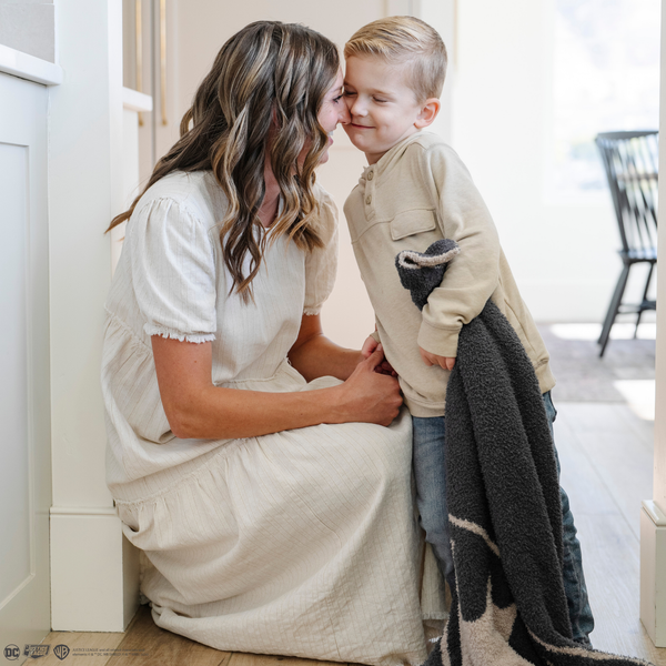 A little boy stands by his mom while holding a black and light gray double-layer Bamboni® blanket with a Batman™ icon on it. The soft blanket is a luxury blanket and a receiving blanket as well as a Saranoni blanket. This kids blanket is perfect for littles. 