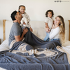 A husband and wife are with their little boy and little girl. They are by a black and light gray double-layer Bamboni® blanket with a Batman™ icon on it. The soft blanket is a luxury blanket and a throw blanket as well as a Saranoni blanket. 