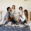 A husband and wife are with their little boy and little girl. They are by a black and light gray double-layer Bamboni® blanket with a Batman™ icon on it. The soft blanket is a luxury blanket and a throw blanket as well as a Saranoni blanket. 