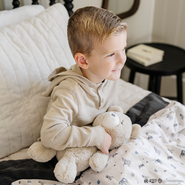 A little boy sits on a bed with a bear stuffed animal and a soft minky blanket featuring a Batman™ design with iconic symbols like the Bat-Signal, Batman logo, and dynamic illustrations of Batman in action. The lightweight white background highlights the blue and black superhero details, making this cozy blanket perfect for DC fans. The soft blanket is a Saranoni blanket. The other side of the blanket is a lush material that is a black/gray.