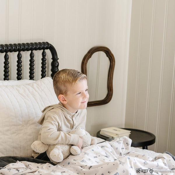 A little boy sits on a bed with a bear stuffed animal and a soft minky blanket featuring a Batman™ design with iconic symbols like the Bat-Signal, Batman logo, and dynamic illustrations of Batman in action. The lightweight white background highlights the blue and black superhero details, making this cozy blanket perfect for DC fans. The soft blanket is a Saranoni blanket. The other side of the blanket is a lush material that is a black/gray.