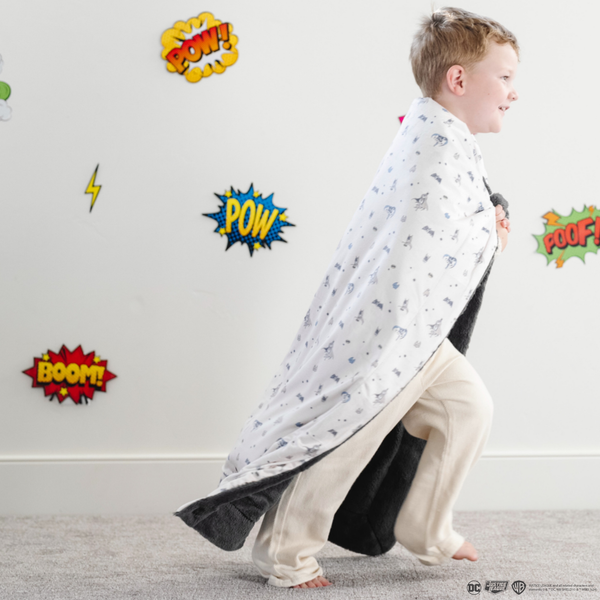 A little boy is wrapped in a soft minky blanket featuring a Batman™ design with iconic symbols like the Bat-Signal, Batman logo, and dynamic illustrations of Batman in action. The lightweight white background highlights the blue and black superhero details, making this cozy blanket perfect for DC fans. The soft blanket is a Saranoni blanket. The other side of the blanket is a lush material that is a black/gray.