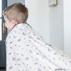 A little boy is wrapped in a soft minky blanket featuring a Batman™ design with iconic symbols like the Bat-Signal, Batman logo, and dynamic illustrations of Batman in action. The lightweight white background highlights the blue and black superhero details, making this cozy blanket perfect for DC fans. The soft blanket is a Saranoni blanket. The other side of the blanket is a lush material that is a black/gray.