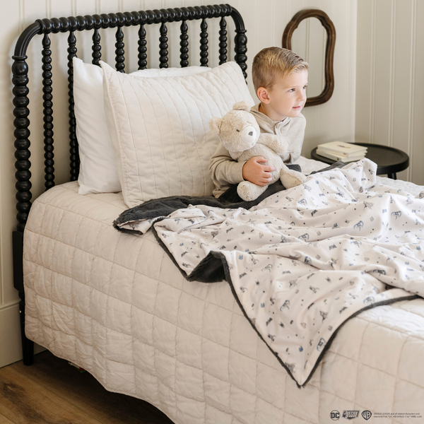 A little boy sits on a bed with a bear stuffed animal and a soft minky blanket featuring a Batman™ design with iconic symbols like the Bat-Signal, Batman logo, and dynamic illustrations of Batman in action. The lightweight white background highlights the blue and black superhero details, making this cozy blanket perfect for DC fans. The soft blanket is a Saranoni blanket. The other side of the blanket is a lush material that is a black/gray.