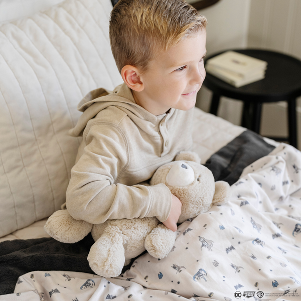 A little boy sits on a bed with a bear stuffed animal and a soft minky blanket featuring a Batman™ design with iconic symbols like the Bat-Signal, Batman logo, and dynamic illustrations of Batman in action. The lightweight white background highlights the blue and black superhero details, making this cozy blanket perfect for DC fans. The soft blanket is a Saranoni blanket. The other side of the blanket is a lush material that is a black/gray.