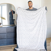 A man holding a soft minky blanket featuring a Batman™ design with iconic symbols like the Bat-Signal, Batman logo, and dynamic illustrations of Batman in action. The lightweight white background highlights the blue and black superhero details, making this cozy blanket perfect for DC fans. The soft blanket is a Saranoni blanket. The other side of the blanket is a lush material that is a black/gray.