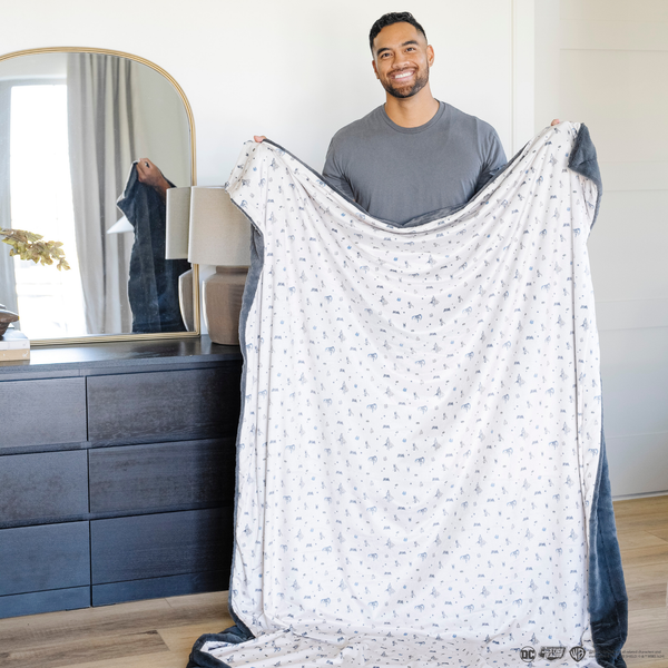 A man holds a soft minky blanket featuring a Batman™ design with iconic symbols like the Bat-Signal, Batman logo, and dynamic illustrations of Batman in action. The lightweight white background highlights the blue and black superhero details, making this cozy blanket perfect for DC fans. The soft blanket is a Saranoni blanket. The other side of the blanket is a lush material that is a black/gray.