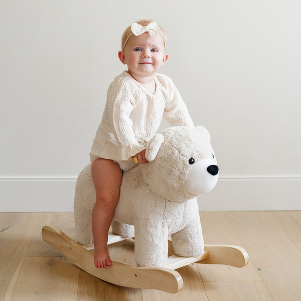 A happy baby girl with a light-colored bow smiles as she rides a soft plush bear rocker, wearing a cozy, cream-colored outfit. This adorable scene captures the essence of toddler playtime and comfort, making it ideal for parents seeking the perfect gifts for their little ones. Featuring plush toys and stylish baby clothing, this image showcases the ultimate in playful nursery decor.