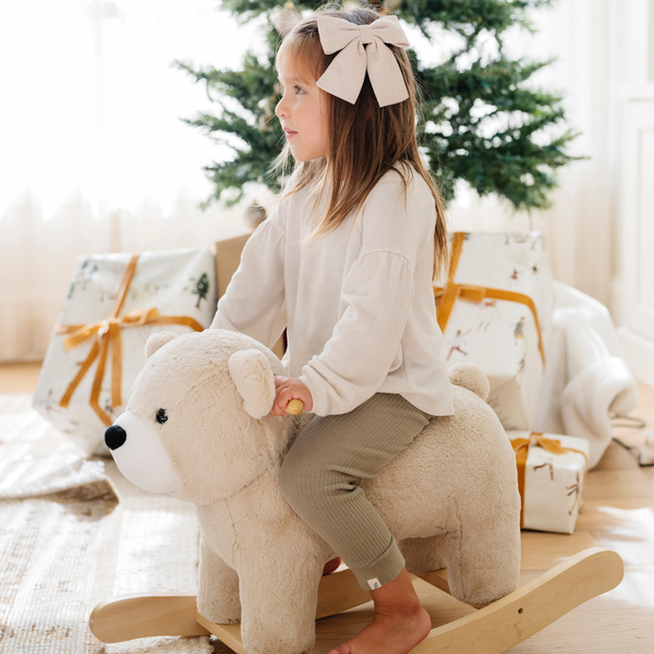 A cheerful girl with long hair and a large bow sits on a plush bear rocker, gazing into the distance. Dressed in a cozy outfit, she is surrounded by beautifully wrapped gifts and a decorated Christmas tree, creating a warm and festive atmosphere. This delightful scene highlights the joy of childhood play and makes for an ideal holiday gift for little ones.