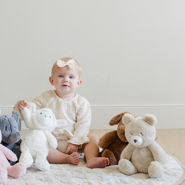 A happy baby sits on a soft quilt surrounded by adorable stuffed animals, including a fluffy lamb and teddy bear. The child, wearing a cute outfit and a bow headband, joyfully holds the plush lamb, showcasing the perfect cuddly toys for infants. This heartwarming scene captures the essence of childhood comfort and the importance of soft toys for imaginative play and nurturing.