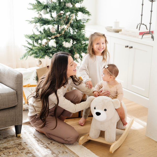 A joyful scene featuring a mother interacting with her two young daughters in a cozy living room adorned with a Christmas tree. The older girl stands beside the plush bear rocker, while the baby girl sits on it, wearing a cute outfit and a bow in her hair. This heartwarming moment captures the essence of family playtime and the joy of childhood, making it perfect for promoting family-friendly products and holiday gift ideas.