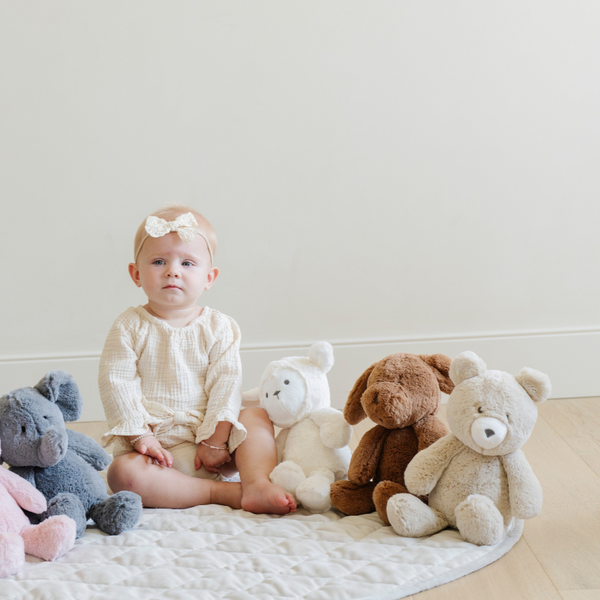 A baby girl sits on a playmat, surrounded by an adorable collection of stuffed animals, including a fluffy lamb, a teddy bear, dog, and a playful elephant. The child, wearing a cute outfit and a bow headband, gazes at the camera while holding the plush lamb, showcasing the perfect cuddly toys for infants. This charming scene highlights the warmth and comfort of soft toys, essential for nurturing imaginative play and childhood joy.