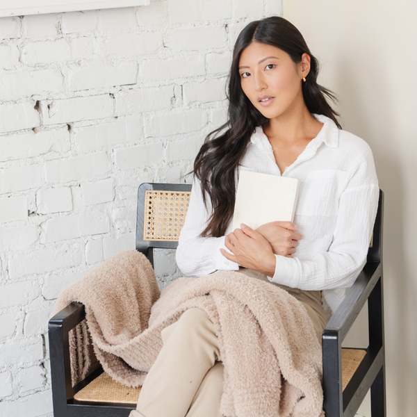 A woman sits with a Bamboni® blanket by Saranoni. This ultra-soft blanket is a light brown blanket and made from a premium blend of bamboo rayon and poly microfiber, offering a lightweight, breathable, and fluffy feel. Perfect as a throw blanket, luxury blanket, plush throw, or soft blanket, it adds warmth and elegance to any living room, bedroom, or home décor setting. Designed for ultimate comfort, this buttery-soft Saranoni blanket is ideal for gifting. Enjoy this cozy blanket for years to come! 