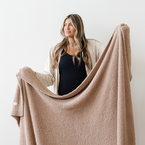 A woman holds up a Bamboni® blanket by Saranoni. This ultra-soft blanket is a light brown blanket and made from a premium blend of bamboo rayon and poly microfiber, offering a lightweight, breathable, and fluffy feel. Perfect as a extra large blanket, luxury blanket, plush blanket, or soft blanket, it adds warmth and elegance to any living room, bedroom, or home décor setting. Designed for ultimate comfort, this buttery-soft Saranoni blanket is ideal for gifting. Enjoy this cozy blanket for years to come! 