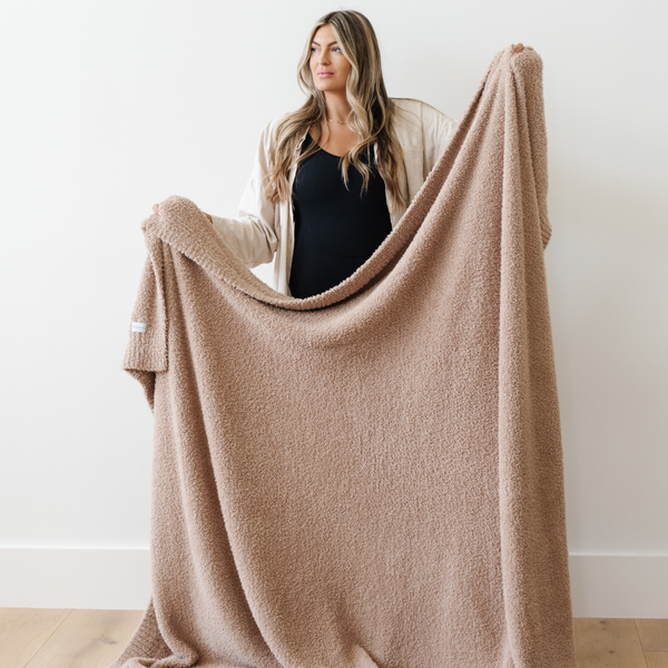 A woman holds up a Bamboni® blanket by Saranoni. This ultra-soft blanket is a light brown blanket and made from a premium blend of bamboo rayon and poly microfiber, offering a lightweight, breathable, and fluffy feel. Perfect as a extra large blanket, luxury blanket, or soft blanket, it adds warmth and elegance to any living room, bedroom, or home décor setting. Designed for ultimate comfort, this buttery-soft Saranoni blanket is ideal for gifting. Enjoy this cozy blanket for years to come!