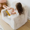 A young girl sits comfortably in a soft white plush chair, engrossed in reading a colorful book. Her legs are casually draped over the armrest, and she wears a charming white dress, radiating a sense of relaxation and joy. This delightful scene showcases the perfect blend of cozy children's furniture and the joy of reading, making it an ideal choice for parents looking to create a nurturing reading nook for their little ones.