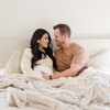 A husband and wife smile and sit with a light tan luxurious patterned faux fur extra large throw blanket that is a Saranoni blanket. This ultra-soft, cozy blanket adds a touch of elegance and warmth to any space, perfect as a soft oversized blanket or fluffy blanket for lounging. Ideal for those seeking a high-quality artificial fur blanket or luxury blanket, this versatile and stylish soft blanket enhances both comfort and decor. Shop the best blankets and throws for ultimate relaxation and cozy vibes.