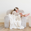 A cheerful girl lounges comfortably on a soft plush chair, wrapped in a cozy, patterned blanket. She smiles as she relaxes, showcasing the inviting texture of the chair and the warm, comforting design of the blanket. This delightful scene highlights the perfect combination of stylish children's furniture and cozy textiles, making it an ideal addition to any child's playroom or reading nook.