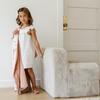 A young girl in a white dress stands beside a plush white chair, holding a cozy blanket with playful designs. She smiles at the camera, exuding warmth and charm in a bright and inviting room. This delightful scene highlights the perfect pairing of stylish children's furniture and soft, comforting blankets, making it an ideal choice for creating a cozy space for kids.