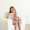 A cheerful girl sits comfortably in a soft white plush chair, smiling as she holds her favorite pink stuffed bunny. Wearing a cute outfit with a bow in her hair, she embodies the joy of childhood. This scene showcases stylish and comfortable children's furniture, perfect for moms searching for cozy seating options and adorable plush toys for their kids' playrooms or reading nooks.