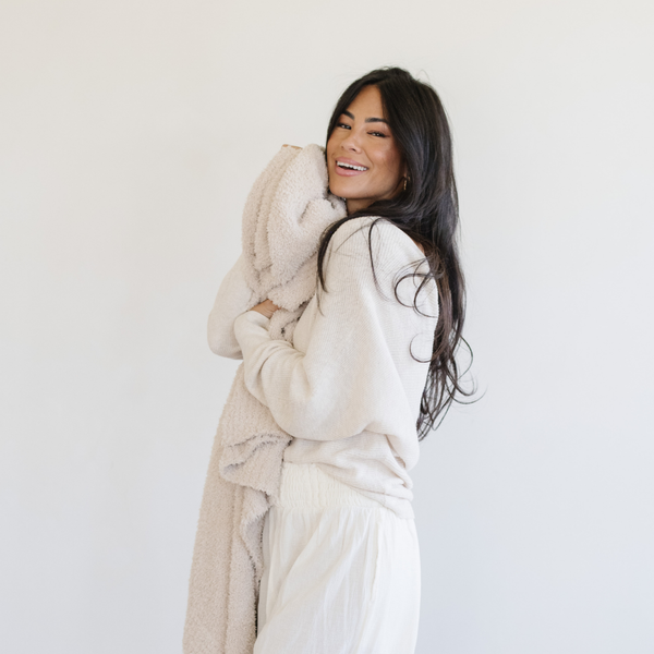 A woman smiles and hugs a Bamboni® blanket by Saranoni. This ultra-soft blanket is a cream blanket and made from a premium blend of bamboo rayon and poly microfiber, offering a lightweight, breathable, and fluffy feel. Perfect as a extra large blanket, luxury blanket, or soft blanket, it adds warmth and elegance to any living room, bedroom, or home décor setting. Designed for ultimate comfort, this buttery-soft Saranoni blanket is ideal for gifting. Enjoy this cozy blanket for years to come!