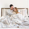 A husband and wife sit under a blue and white Saranoni Queen/King muslin quilt in a soft stripe print. This luxury bedding blanket features four layers of ultra-soft bamboo muslin, creating an airy yet cozy feel. Designed with a stitched quilted pattern and a one-inch border, this breathable blanket is perfect for muslin lovers looking for extra warmth and comfort. This luxury blanket is the softest blanket and a Saranoni blanket. 