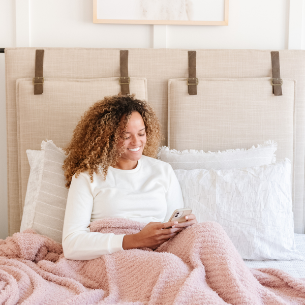 A woman smiles sits with a luxurious ribbed Bamboni® blanket by Saranoni, the blanket is light pink. This ultra-soft blanket is crafted from bamboo rayon and poly microfiber, making it lightweight, breathable, and fluffy. Ideal as a cozy blanket, throw blanket, luxury blanket, soft blanket, or stylish home accent, this Saranoni blanket brings warmth, comfort, and timeless elegance to any living room or bedroom.