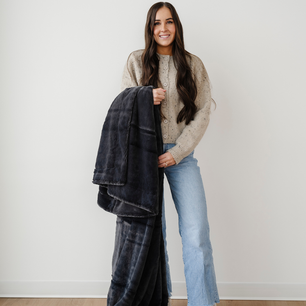 A woman smiles and holds a charcoal blanket that is a lush blanket with a luxurious texture. This cozy blanket is great for gifting, relaxing, or as an addition to your home decor. This soft blanket is a luxury blanket and a Saranoni blanket. Enjoy luxury that lasts.