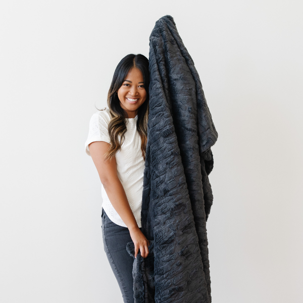 A woman smiles and holds a charcoal luxurious patterned faux fur throw blanket that is a Saranoni blanket. This ultra-soft, plush throw blanket adds a touch of elegance and warmth to any space, perfect as a soft throw blanket or fluffy blanket for lounging. Ideal for those seeking a high-quality artificial fur blanket or luxury blanket, this versatile and stylish soft blanket enhances both comfort and decor. Shop the best blankets and throws for ultimate relaxation and cozy vibes.