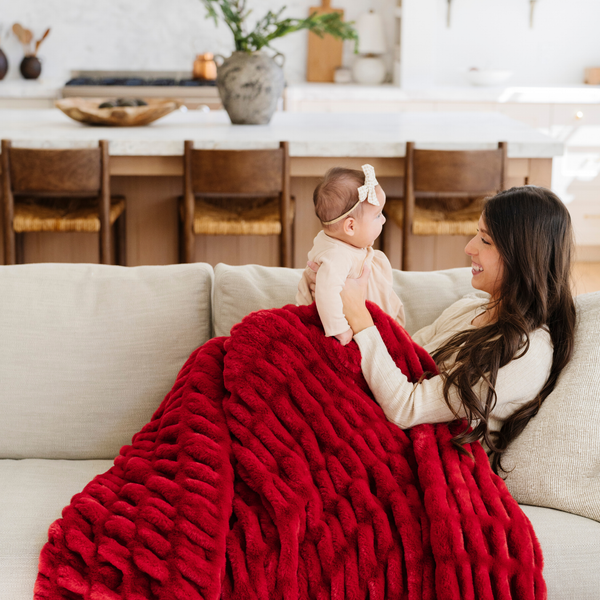 Mother and baby sharing a cozy moment on the sofa with a plush red double ruched blanket, adding warmth and holiday spirit to the room. Ideal for festive decor and family relaxation. The soft blanket is a luxury blanket and a Saranoni blanket.