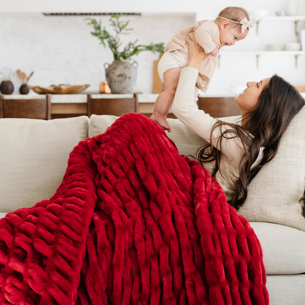 Mother and baby sharing a cozy moment on the sofa with a plush red double ruched blanket, adding warmth and holiday spirit to the room. Ideal for festive decor and family relaxation. The soft blanket is a luxury blanket and a Saranoni blanket.