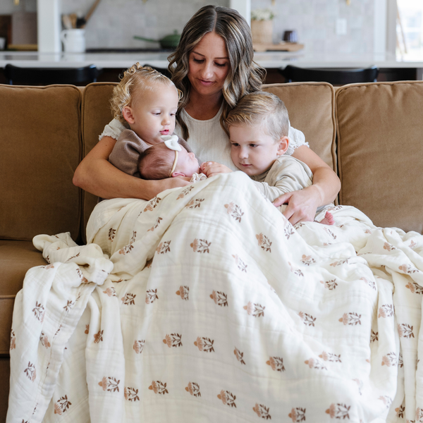 A mother and her three little kids sit with a Saranoni XL muslin quilt in a soft floral print. This luxury blanket features four layers of ultra-soft bamboo muslin, creating an airy yet cozy feel. Designed with a stitched quilted pattern and a one-inch border, this breathable blanket is perfect for muslin lovers looking for extra warmth and comfort. Made from bamboo fabric, this soft muslin quilt is a lightweight quilt that offers a cozy, neutral home décor accent. 