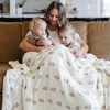 A mother and her three little kids sit with a Saranoni XL muslin quilt in a soft floral print. This luxury blanket features four layers of ultra-soft bamboo muslin, creating an airy yet cozy feel. Designed with a stitched quilted pattern and a one-inch border, this breathable blanket is perfect for muslin lovers looking for extra warmth and comfort. Made from bamboo fabric, this soft muslin quilt is a lightweight quilt that offers a cozy, neutral home décor accent. 