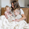 A mother and her three kids sit with a  Saranoni XL muslin quilt in a soft floral print. This luxury blanket features four layers of ultra-soft bamboo muslin, creating an airy yet cozy feel. Designed with a stitched quilted pattern and a one-inch border, this breathable blanket is perfect for muslin lovers looking for extra warmth and comfort. Made from bamboo fabric, this soft muslin quilt is a lightweight quilt that offers a cozy, neutral home décor accent. The softest blanket and a Saranoni blanket.