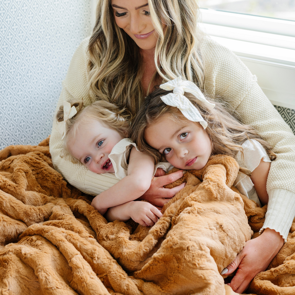 A mother, her two daughters, and a copper colored luxurious patterned faux fur extra large throw blanket that is a Saranoni blanket. This ultra-soft, cozy blanket adds a touch of elegance and warmth to any space, perfect as a soft oversized blanket or fluffy blanket for lounging. Ideal for those seeking a high-quality artificial fur blanket or luxury blanket, this versatile and stylish soft blanket enhances both comfort and decor. Shop the best blankets and throws for ultimate relaxation.