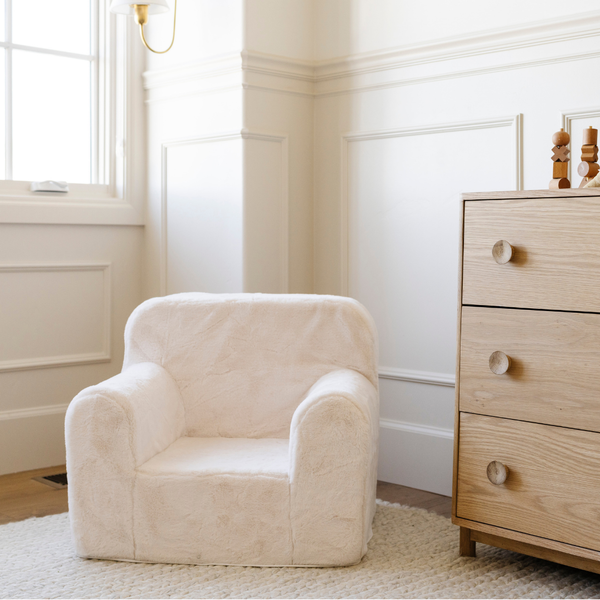 A cozy white plush chair is elegantly placed in a well-lit room with soft natural light streaming through a window. The chair features rounded arms and a plush design, making it an inviting spot for children to relax. Nearby, a wooden dresser adds a touch of warmth to the decor, showcasing a perfect blend of style and comfort for a child's play area.