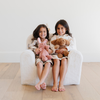 Two joyful little girls sit side by side on a plush white chair, each holding their favorite stuffed animals. The girl on the left cuddles a soft pink bunny, while the girl on the right holds a brown plush dog. This charming scene highlights the perfect combination of cozy kids' furniture and beloved stuffed toys, making it an ideal choice for parents seeking stylish and comfortable seating for children.