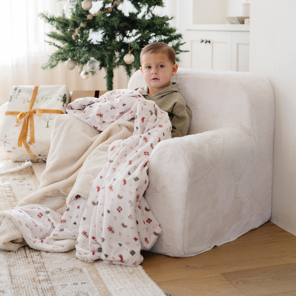 A young boy sits comfortably on a plush white chair, wrapped in a cozy blanket adorned with festive designs. He gazes thoughtfully at the camera, surrounded by a warm and inviting room decorated for the holidays, complete with a Christmas tree and beautifully wrapped gifts. This heartwarming scene highlights the perfect combination of kids' furniture and snug blankets, making it an ideal choice for parents looking to create cozy play areas for their children.