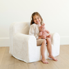 A cheerful girl with long hair sits comfortably in a plush white chair, smiling brightly while holding a soft pink bunny toy. Dressed in a stylish outfit, she radiates joy in a bright and airy room. This delightful scene captures the essence of cozy playtime and is perfect for promoting children's furniture and plush toys.