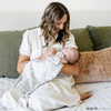 A mother holds her baby girl in a light gray muslin blanket featuring subtle tonal prints of iconic DC superhero logos, including Batman™, Superman™, and Wonder Woman™. The sophisticated neutral design is paired with a soft white plush interior, offering a perfect blend of style and comfort. Perfect for snuggling, showcasing fandom, or elevating your home decor with understated superhero flair. The soft blanket is a Saranoni blanket.