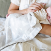 A mother holds her baby girl in a light gray muslin blanket featuring subtle tonal prints of iconic DC superhero logos, including Batman™, Superman™, and Wonder Woman™. The sophisticated neutral design is paired with a soft white plush interior, offering a perfect blend of style and comfort. Perfect for snuggling, showcasing fandom, or elevating your home decor with understated superhero flair. The soft blanket is a Saranoni blanket. 