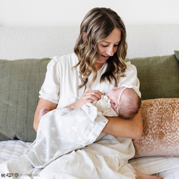 A mother holds her baby girl in a light gray muslin blanket featuring subtle tonal prints of iconic DC superhero logos, including Batman™, Superman™, and Wonder Woman™. The sophisticated neutral design is paired with a soft white plush interior, offering a perfect blend of style and comfort. Perfect for snuggling, showcasing fandom, or elevating your home decor with understated superhero flair. The soft blanket is a Saranoni blanket.