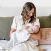 A mother holds her baby girl in a light gray muslin blanket featuring subtle tonal prints of iconic DC superhero logos, including Batman™, Superman™, and Wonder Woman™. The sophisticated neutral design is paired with a soft white plush interior, offering a perfect blend of style and comfort. Perfect for snuggling, showcasing fandom, or elevating your home decor with understated superhero flair. The soft blanket is a Saranoni blanket.