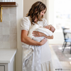 A mother holds her baby girl in a light gray muslin blanket featuring subtle tonal prints of iconic DC superhero logos, including Batman™, Superman™, and Wonder Woman™. The sophisticated neutral design is paired with a soft white plush interior, offering a perfect blend of style and comfort. Perfect for snuggling, showcasing fandom, or elevating your home decor with understated superhero flair. The soft blanket is a Saranoni blanket.