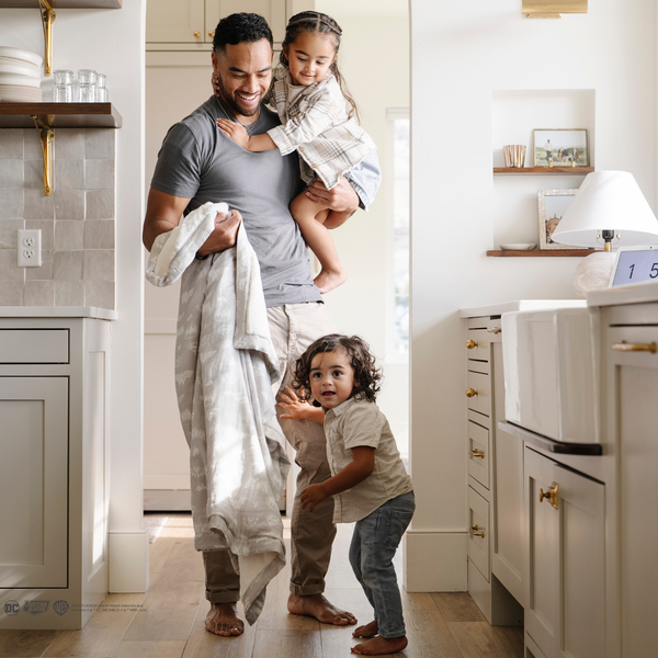 A father stands by his little boy and is holding his little girl and a light gray muslin blanket featuring subtle tonal prints of iconic DC superhero logos, including Batman™, Superman™, and Wonder Woman™. The sophisticated neutral design is paired with a soft white plush interior, offering a perfect blend of style and comfort. Perfect for snuggling, showcasing fandom, or elevating your home decor with understated superhero flair. The soft blanket is a Saranoni blanket.