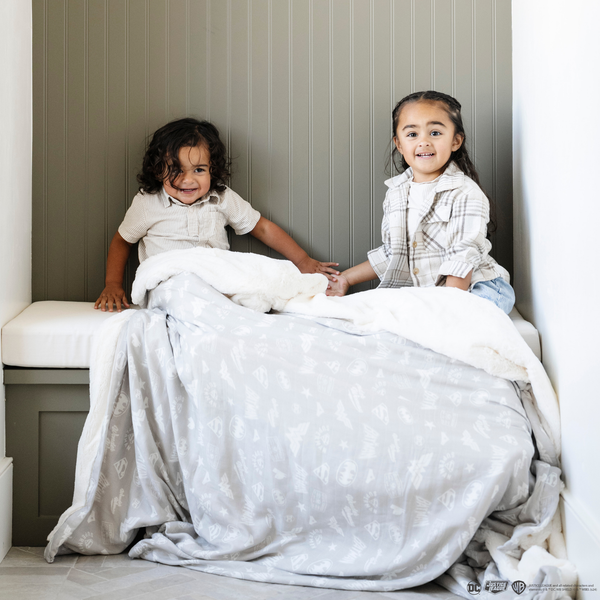 A brother and sister sit under a light gray muslin blanket featuring subtle tonal prints of iconic DC superhero logos, including Batman™, Superman™, and Wonder Woman™. The sophisticated neutral design is paired with a soft white plush interior, offering a perfect blend of style and comfort. Perfect for snuggling, showcasing fandom, or elevating your home decor with understated superhero flair. The soft blanket is a Saranoni blanket.